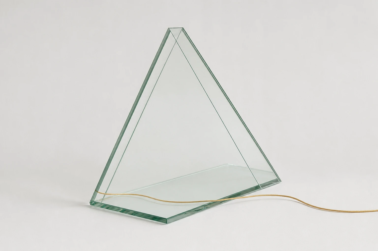 A minimalist studio photograph of a triangular glass sculptural object with a thin brass cord trailing across a soft neutral surface.