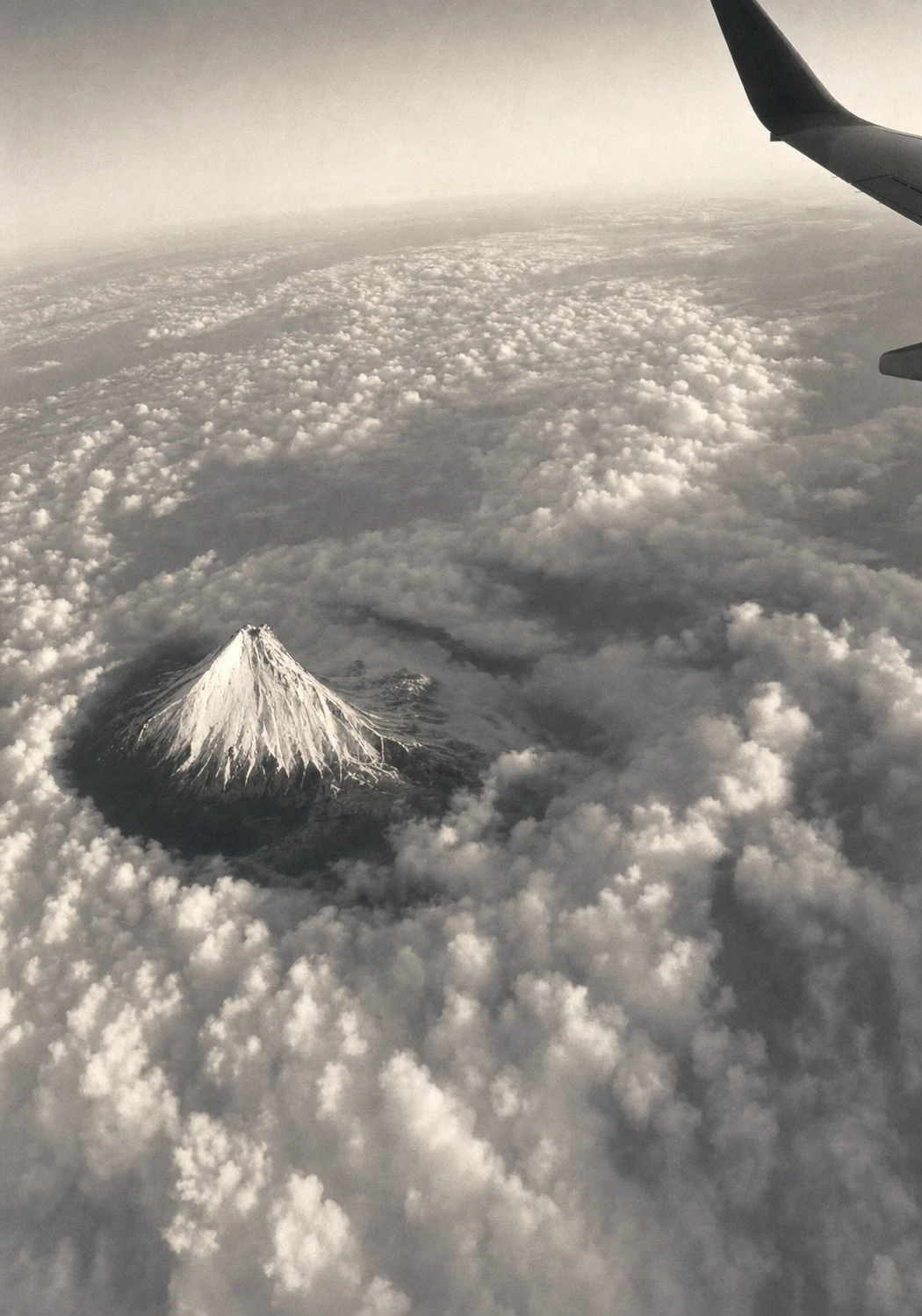 A monochrome aerial photograph of a lone snow-capped conical mountain piercing a sea of clouds, seen from an airplane window with the wingtip visible.