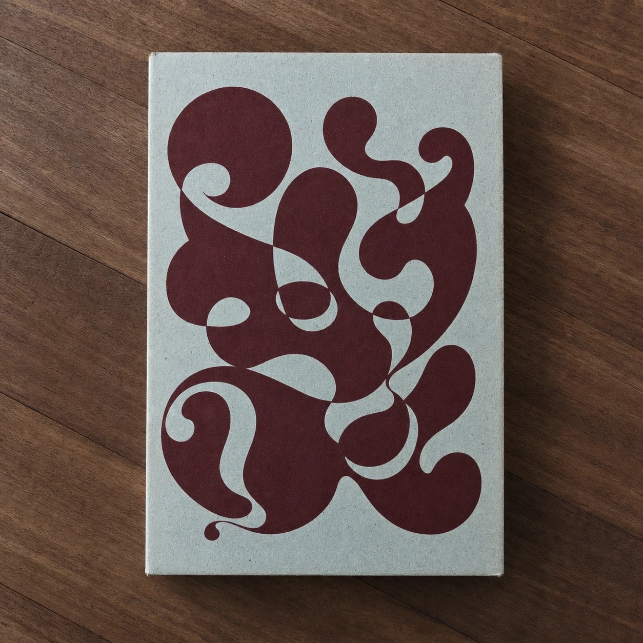 A grey cardboard book or box cover printed with an abstract maroon liquid-organic graphic, photographed flat on a dark wooden surface.