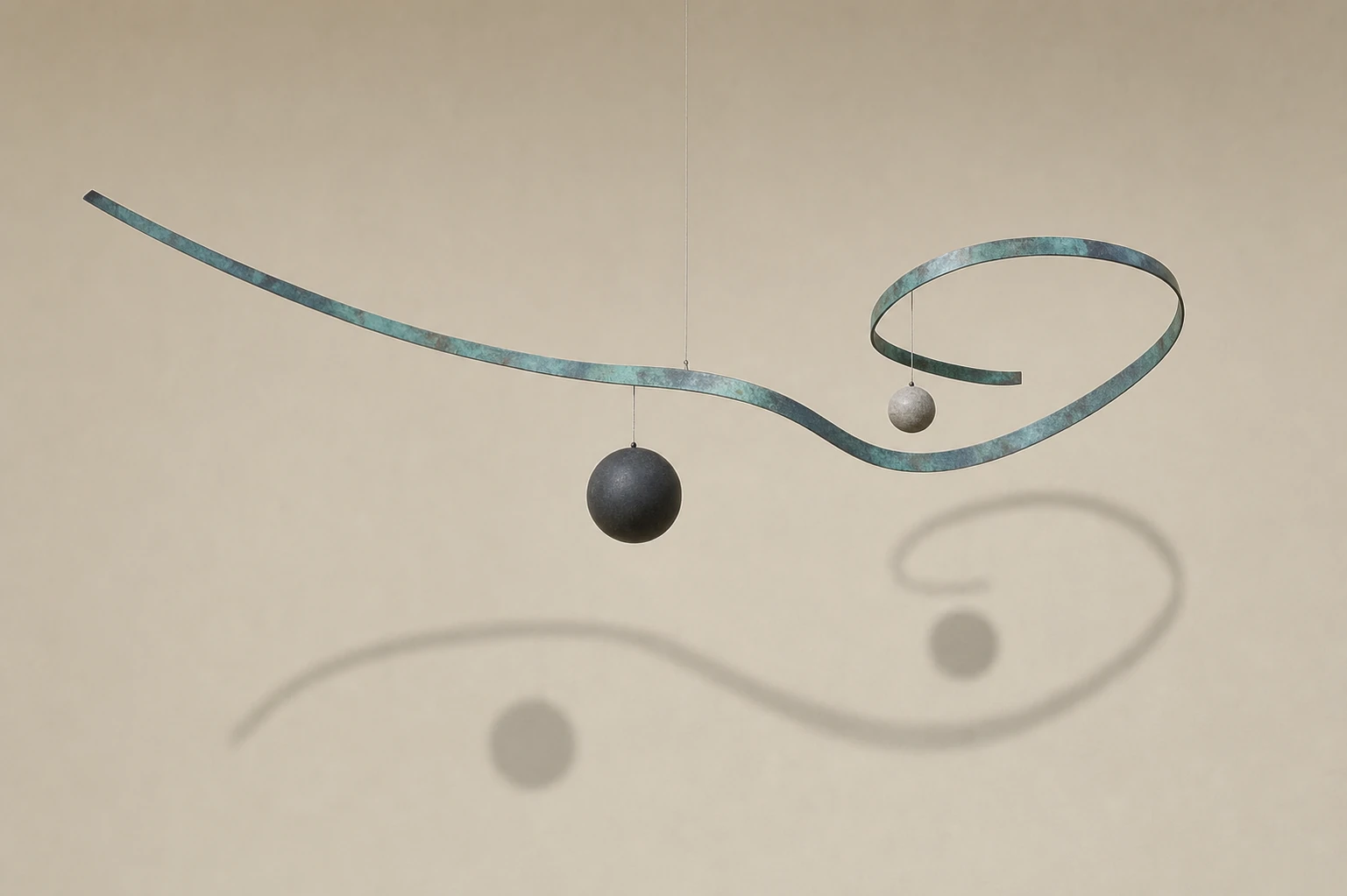 A suspended kinetic sculpture made of a curling patinated metal ribbon and hanging spheres, casting a soft shadow on a warm neutral wall.