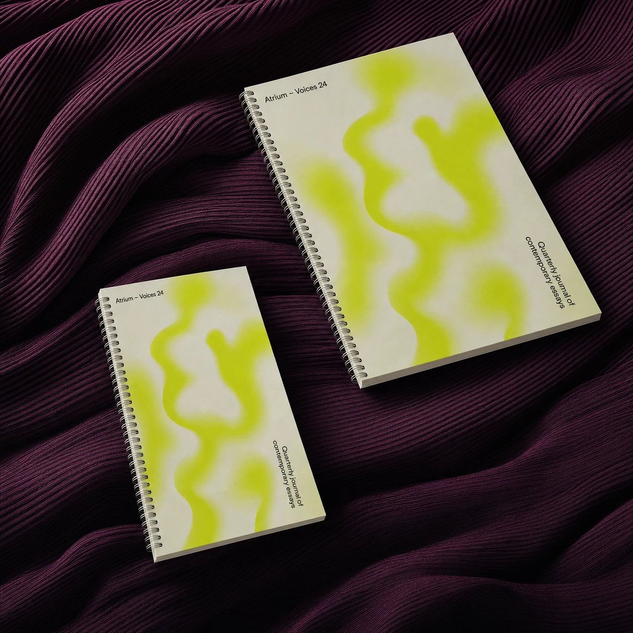 Two spiral-bound journals with soft abstract chartreuse cover art, staged on rich draped burgundy ribbed fabric.