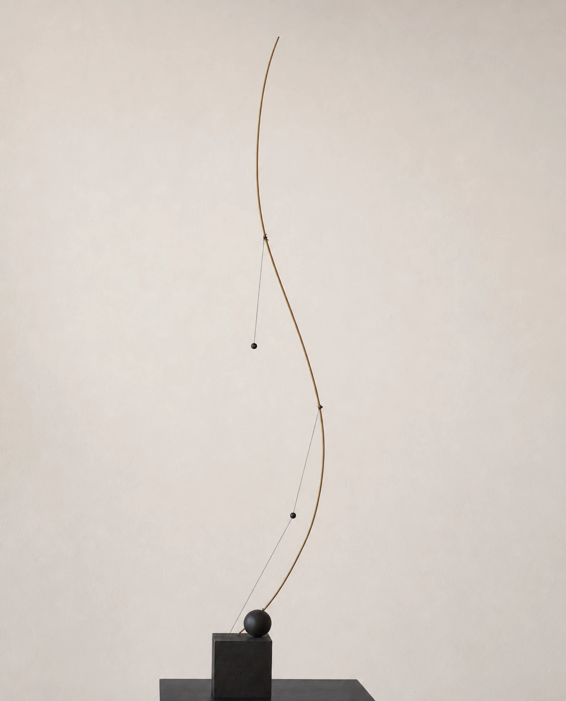 A minimalist kinetic sculpture of a tall curving brass rod and small dark spheres balanced on a dark cubic base, photographed against a soft neutral wall.