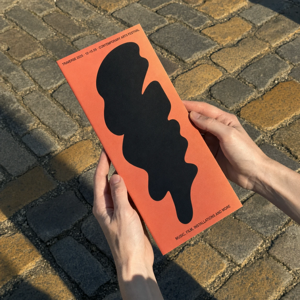 A pair of hands holds a tall orange festival flyer featuring a bold abstract black blob, photographed over a sunlit cobblestone street.