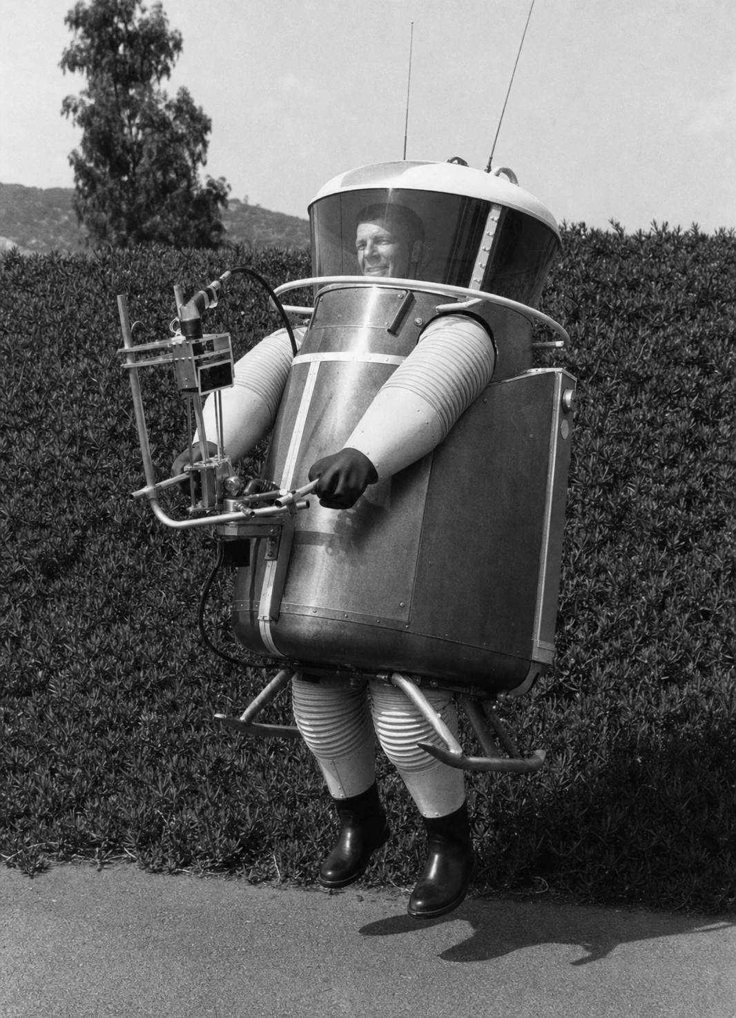 A vintage black-and-white photograph of a smiling man wearing an elaborate retro-futuristic, barrel-shaped pressurized exploration suit standing outdoors.