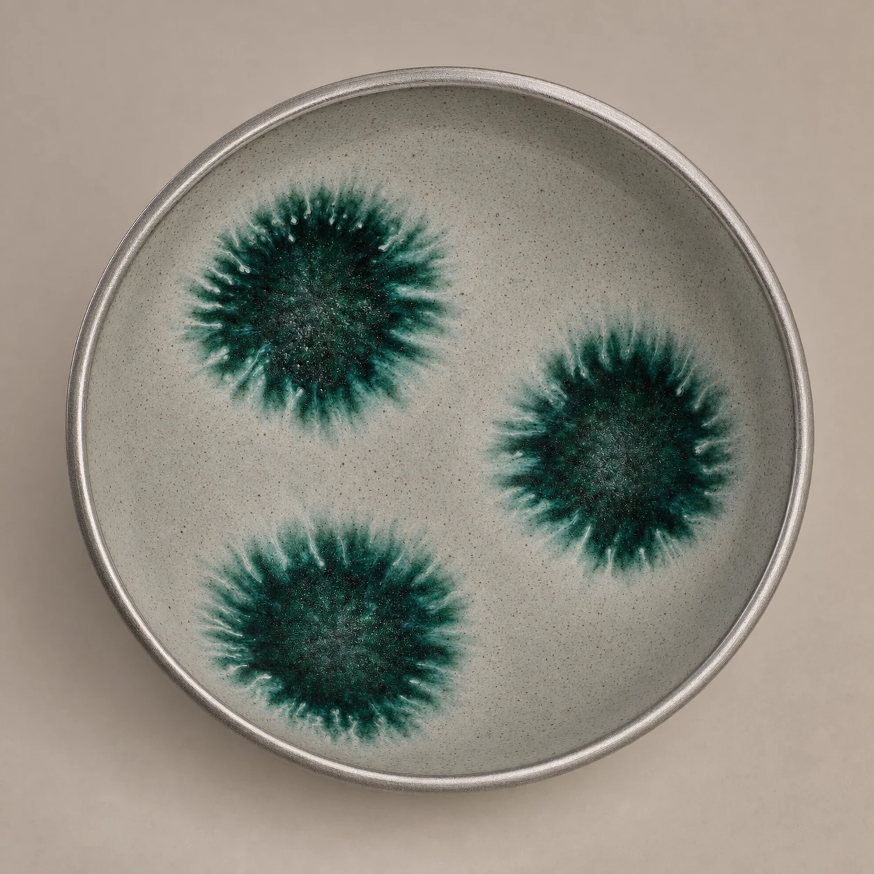 An overhead studio shot of a handmade ceramic bowl with a speckled gray glaze and three burst-like green glaze blooms.