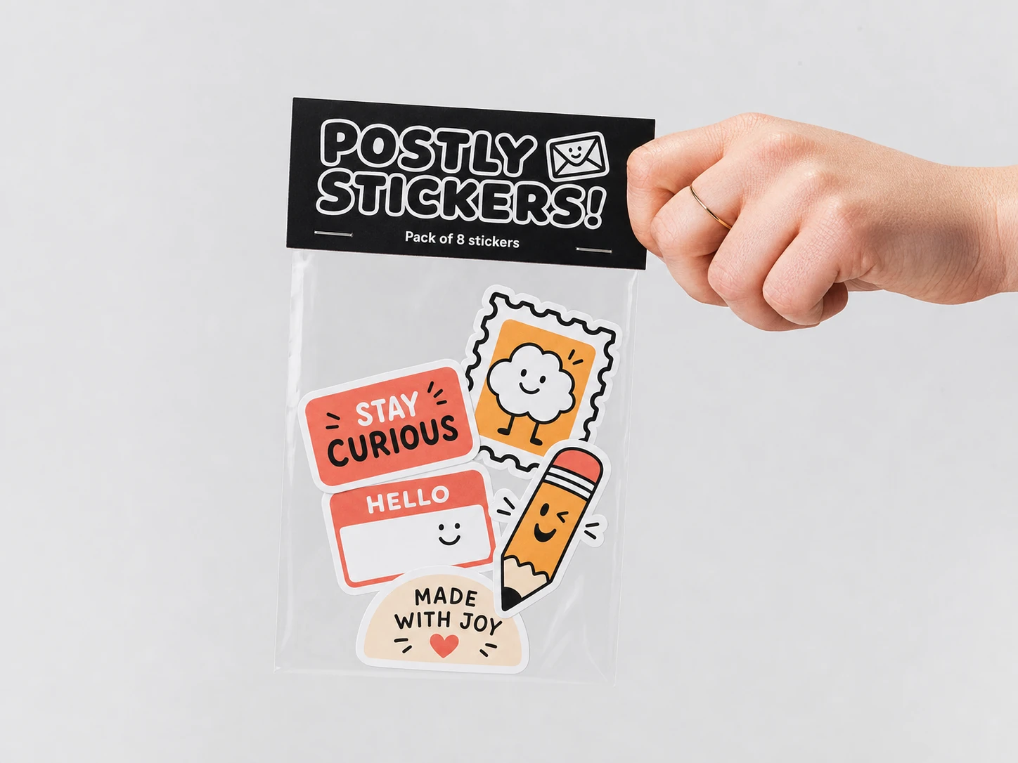 A hand holds a clear sticker pack with a black header card and a set of cute cartoon stickers against a clean light-gray studio background.