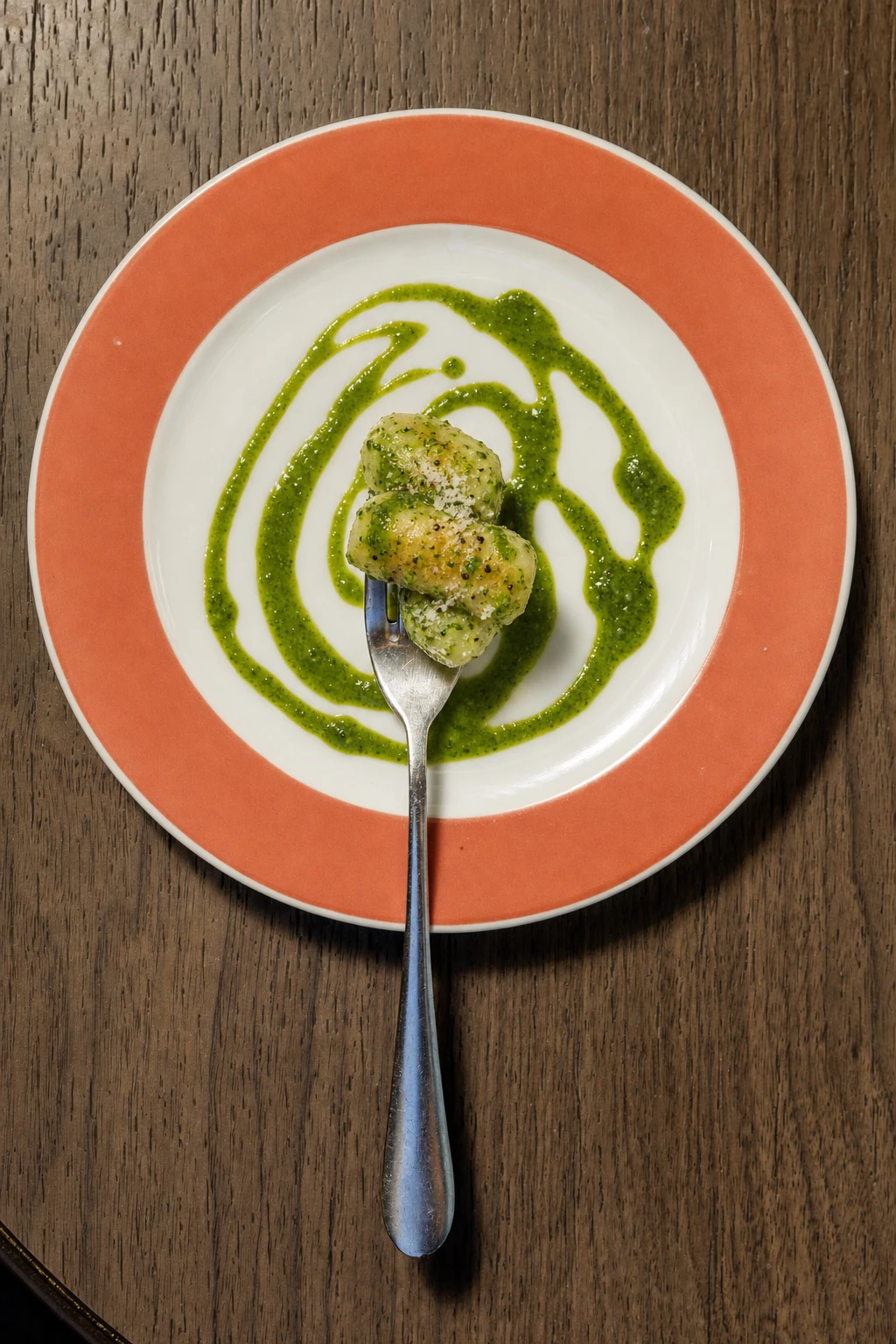 An overhead food photograph of a minimalist plated gnocchi dish with vibrant green pesto on a white plate with a colored rim, set on a dark wood table.