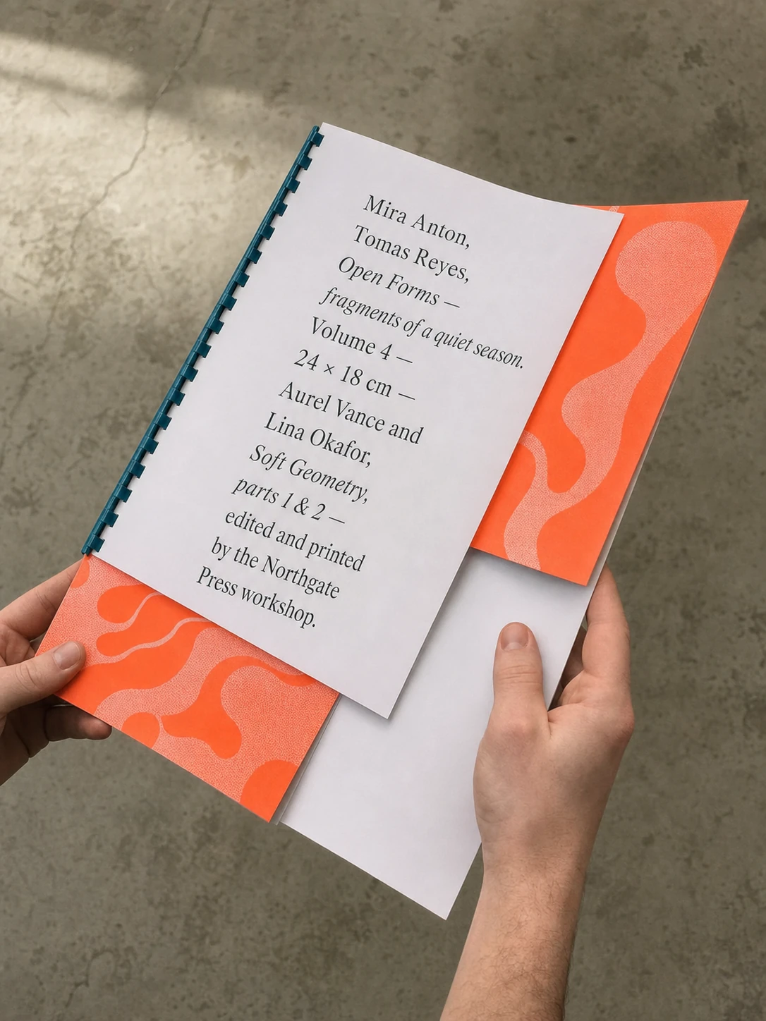 A pair of hands holds a spiral-bound printed booklet with a white centered-text page and bright orange organic-pattern covers over a concrete floor.