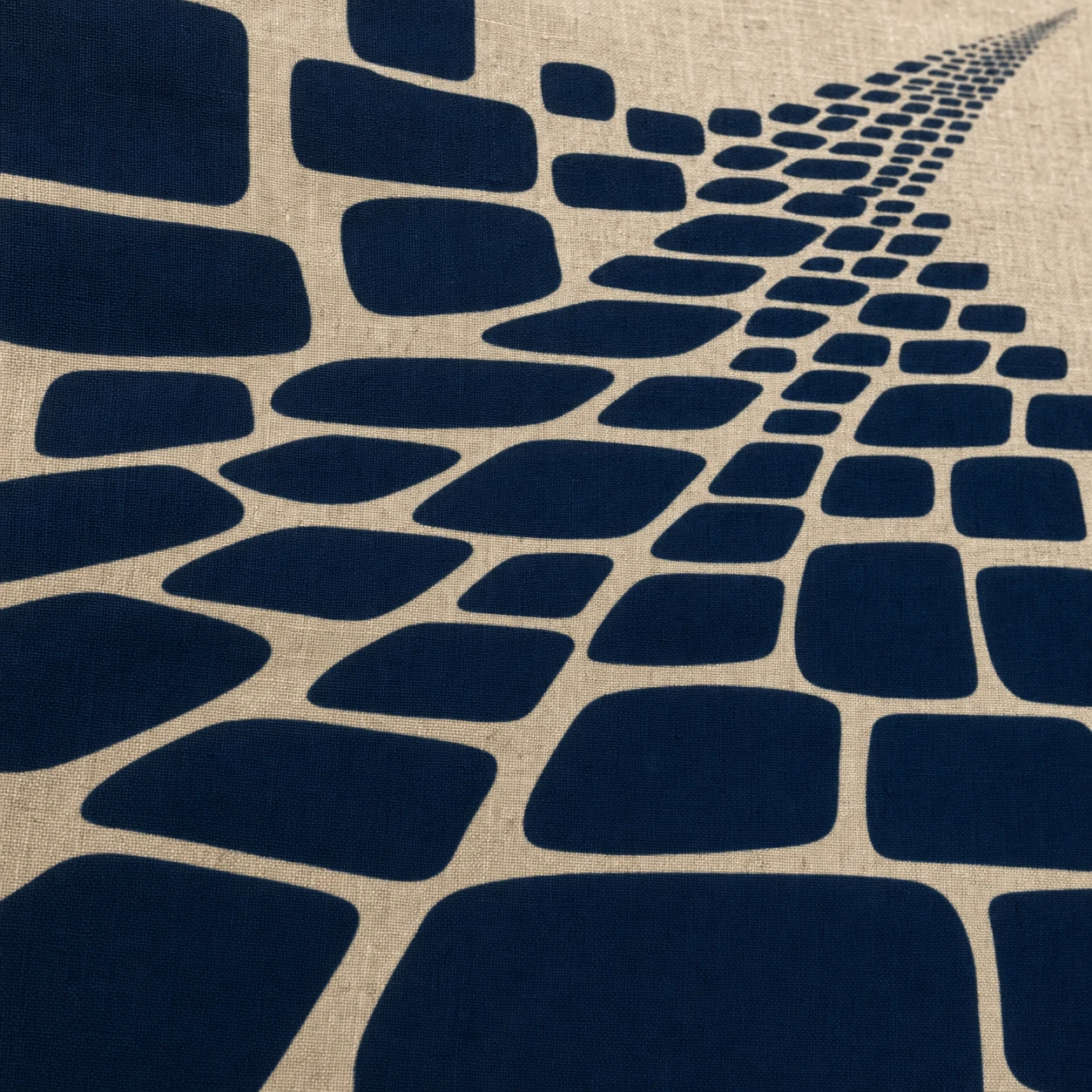 A close-up of natural linen fabric printed with a navy mosaic of organic rounded tiles that scale from large in the foreground to a tiny vanishing trail recedin