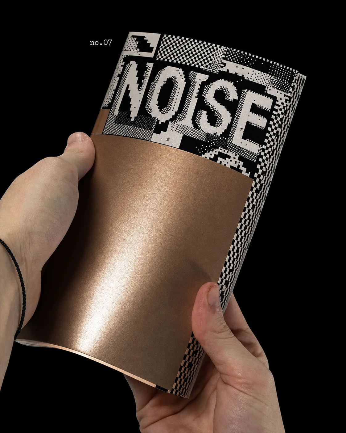 Two hands hold an open metallic-bronze magazine or zine featuring bold pixelated black-and-white cover graphics against a dark backdrop.