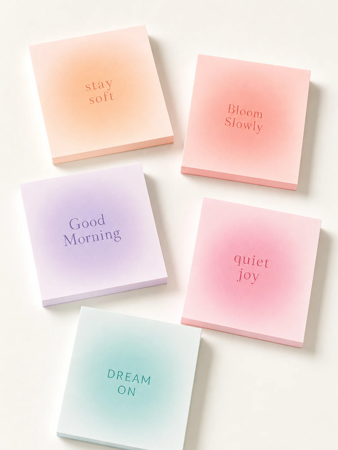 A flat-lay arrangement of five square pastel sticky-note pads, each printed with a short affirmation phrase against a soft radial gradient.