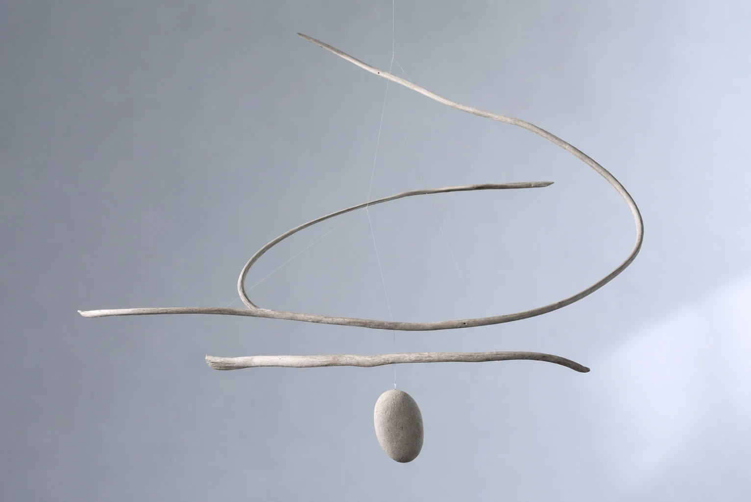 A minimalist hanging mobile made of curved driftwood branches balanced by a single smooth stone, suspended against a soft gray gradient.