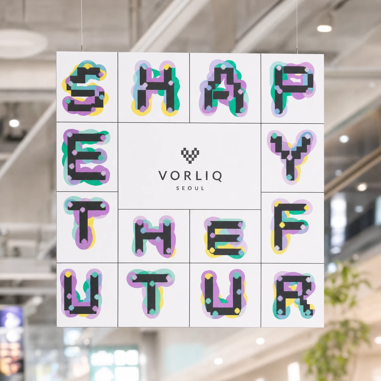 A suspended square signage panel built from a grid of white tiles spelling a bold typographic slogan, with a centered brand lockup, hung in a bright modern inte