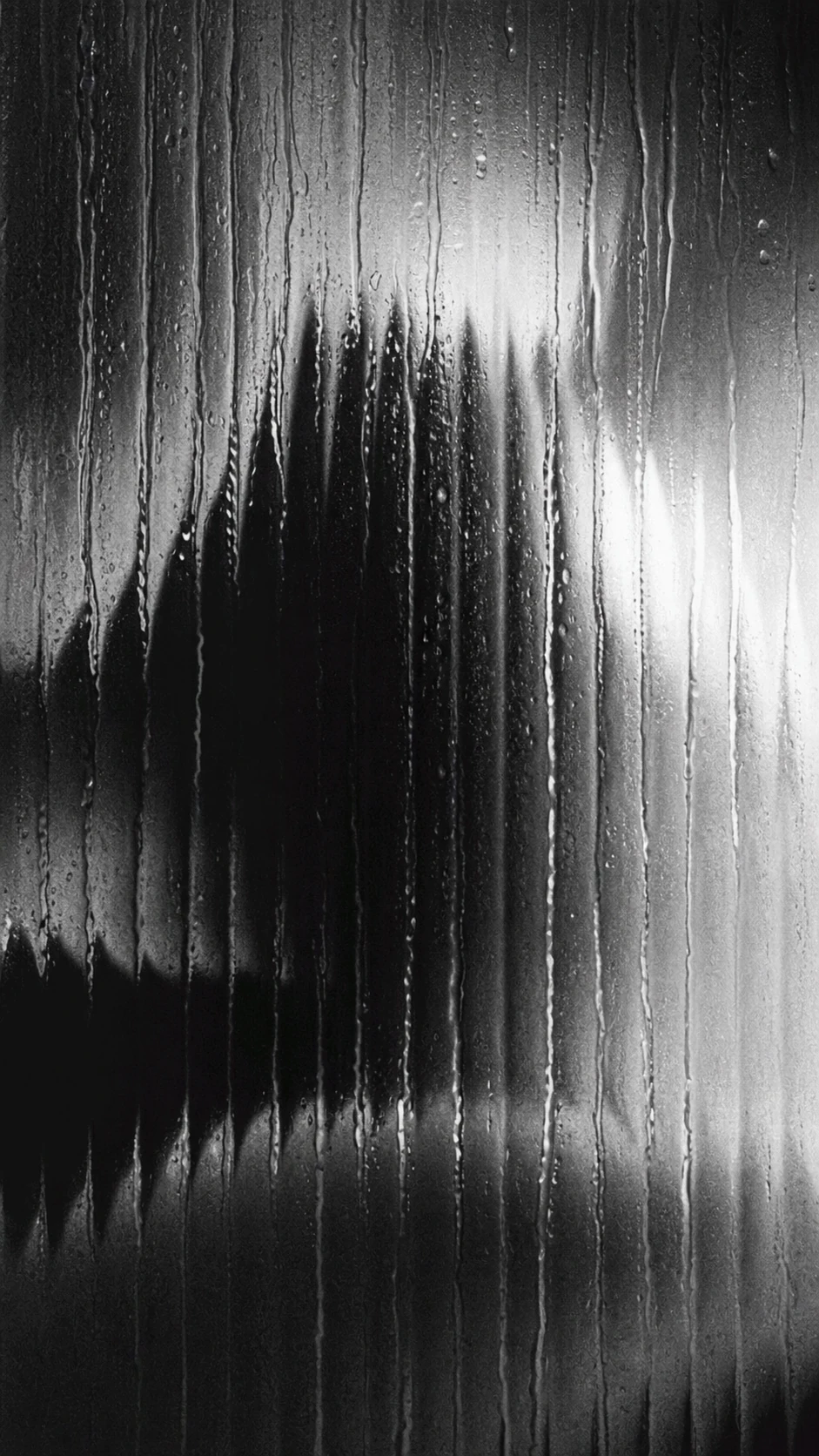 A moody black-and-white image of a blurred human silhouette seen through fogged, water-streaked glass.