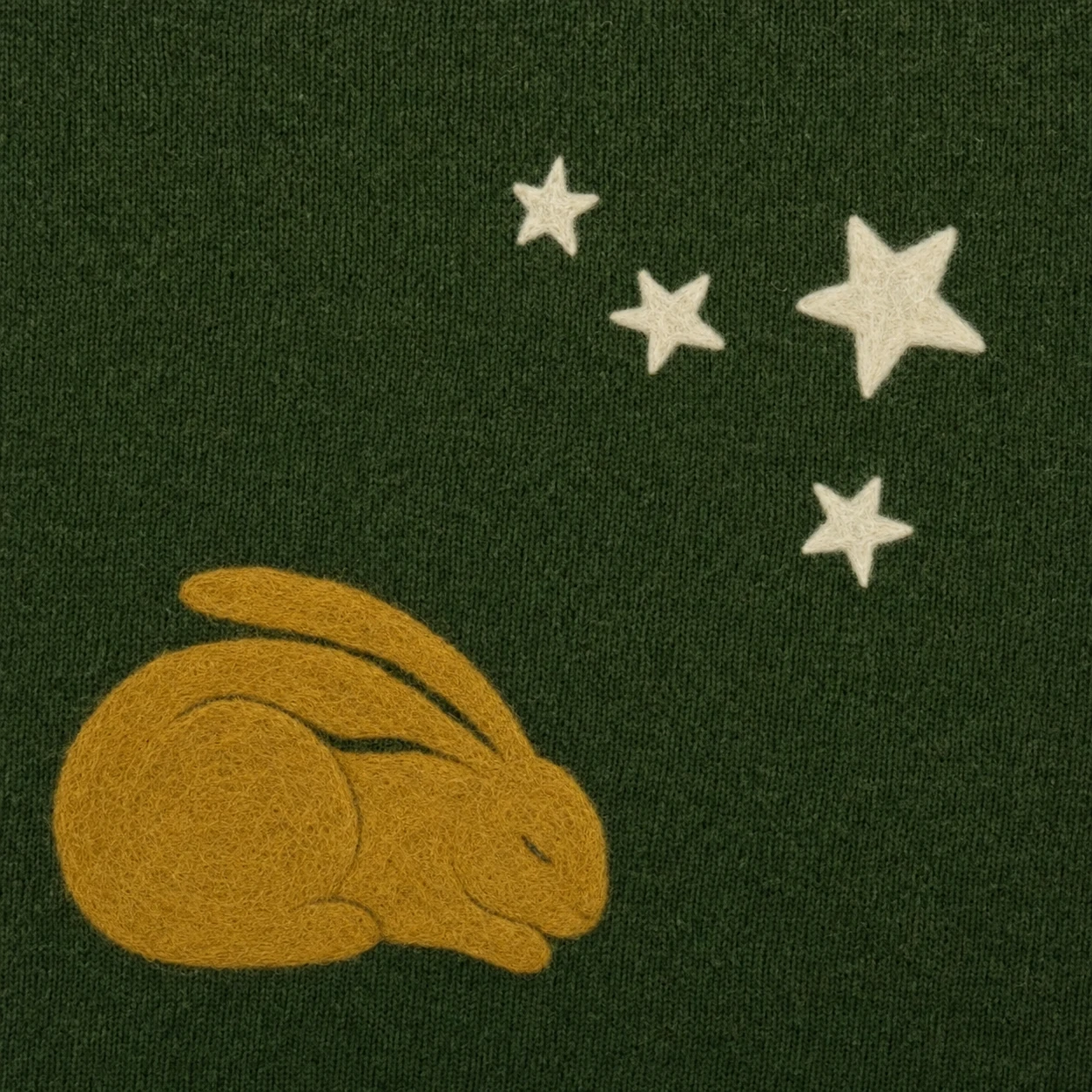 A needle-felted wool illustration of a sleeping golden rabbit beneath a scatter of cream stars on a forest-green knit fabric.
