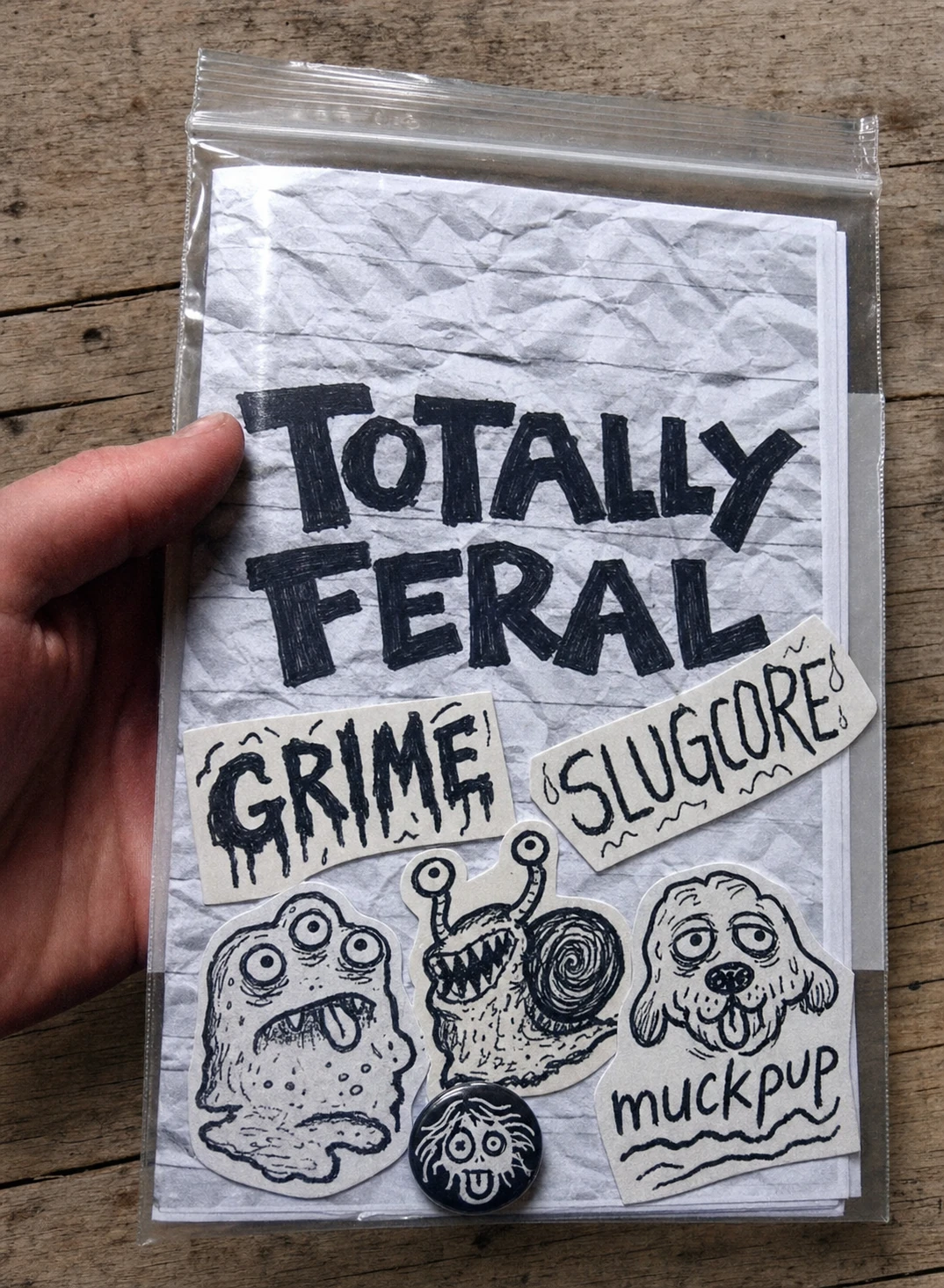 A hand-held DIY zine in a plastic baggie featuring hand-lettered punk titles and ink-drawn monster stickers on crumpled paper, photographed against weathered wo