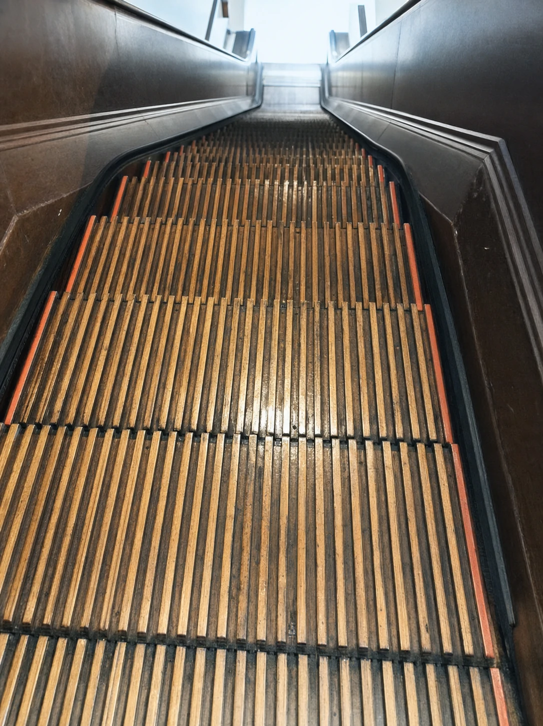 A low-angle photograph looking up a vintage wooden-slat escalator with metal side panels receding toward a bright top.