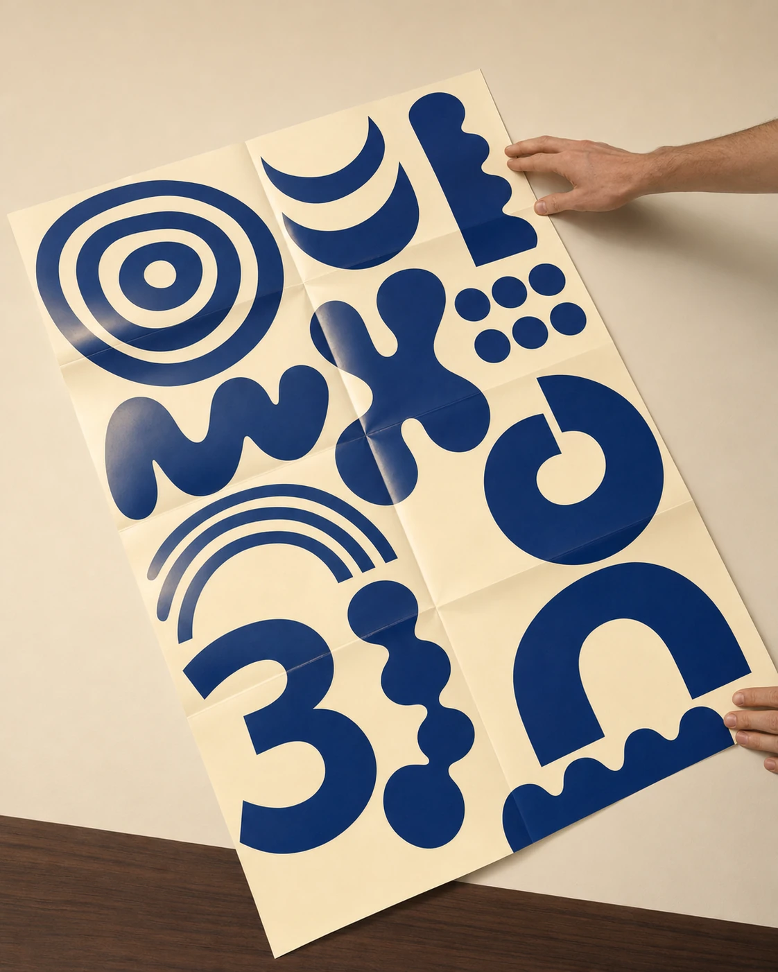 A folded cream-colored poster covered in bold cobalt-blue abstract organic shapes, held up by hands against a neutral wall.