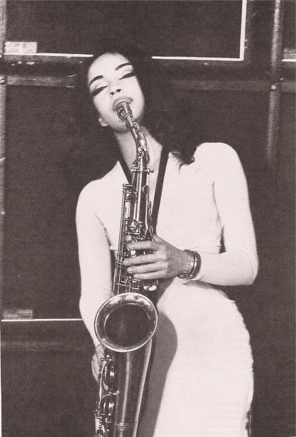 A grainy black-and-white photograph of a woman in a fitted light dress playing a tenor saxophone on a dim stage.