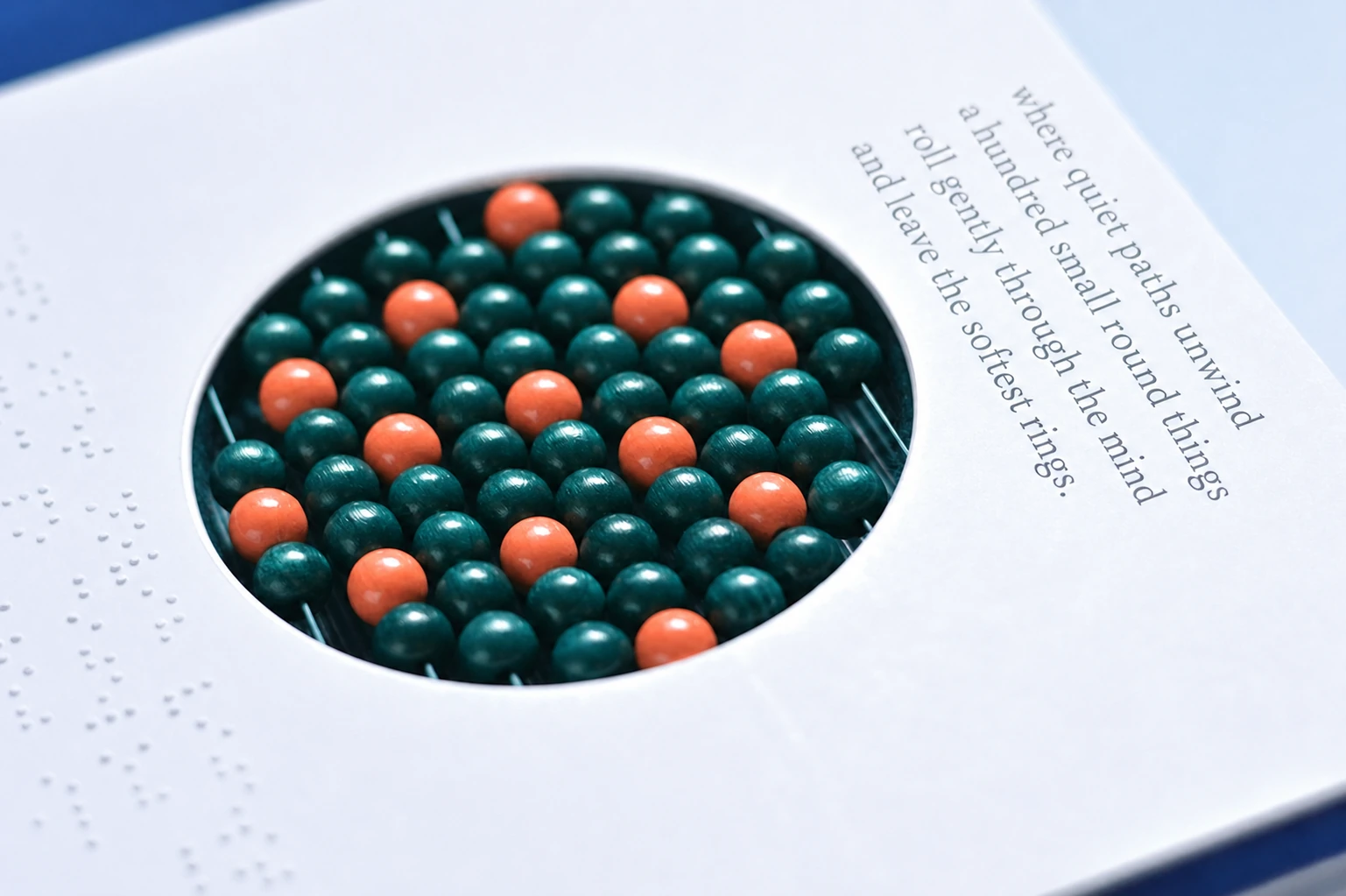 A macro photograph of a tactile abacus-like grid of glossy wooden beads viewed through a circular die-cut window in a white card, accompanied by embossed braill
