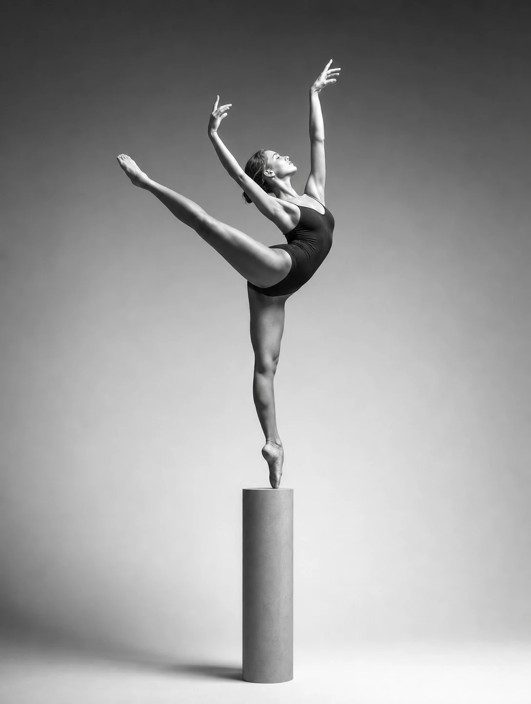 A black-and-white studio photograph of a ballet dancer balanced en pointe atop a tall cylindrical pedestal in an extended arabesque.