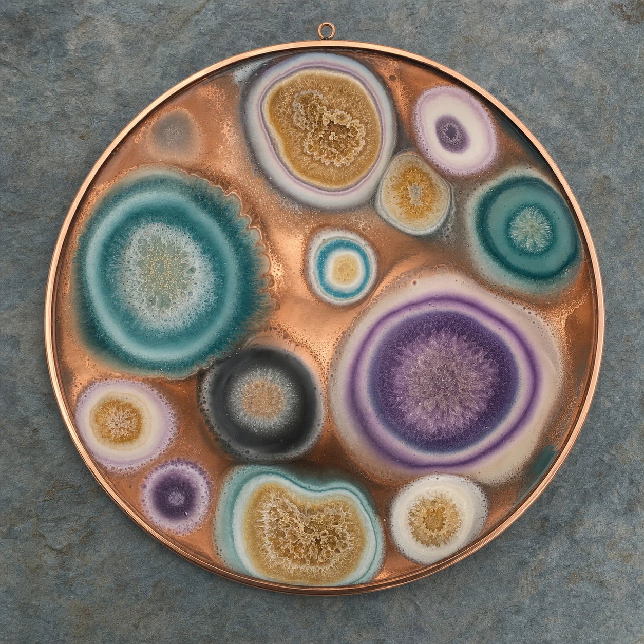 A circular copper enamel wall plaque with abstract organic cellular bursts in jewel tones, mounted on slate.