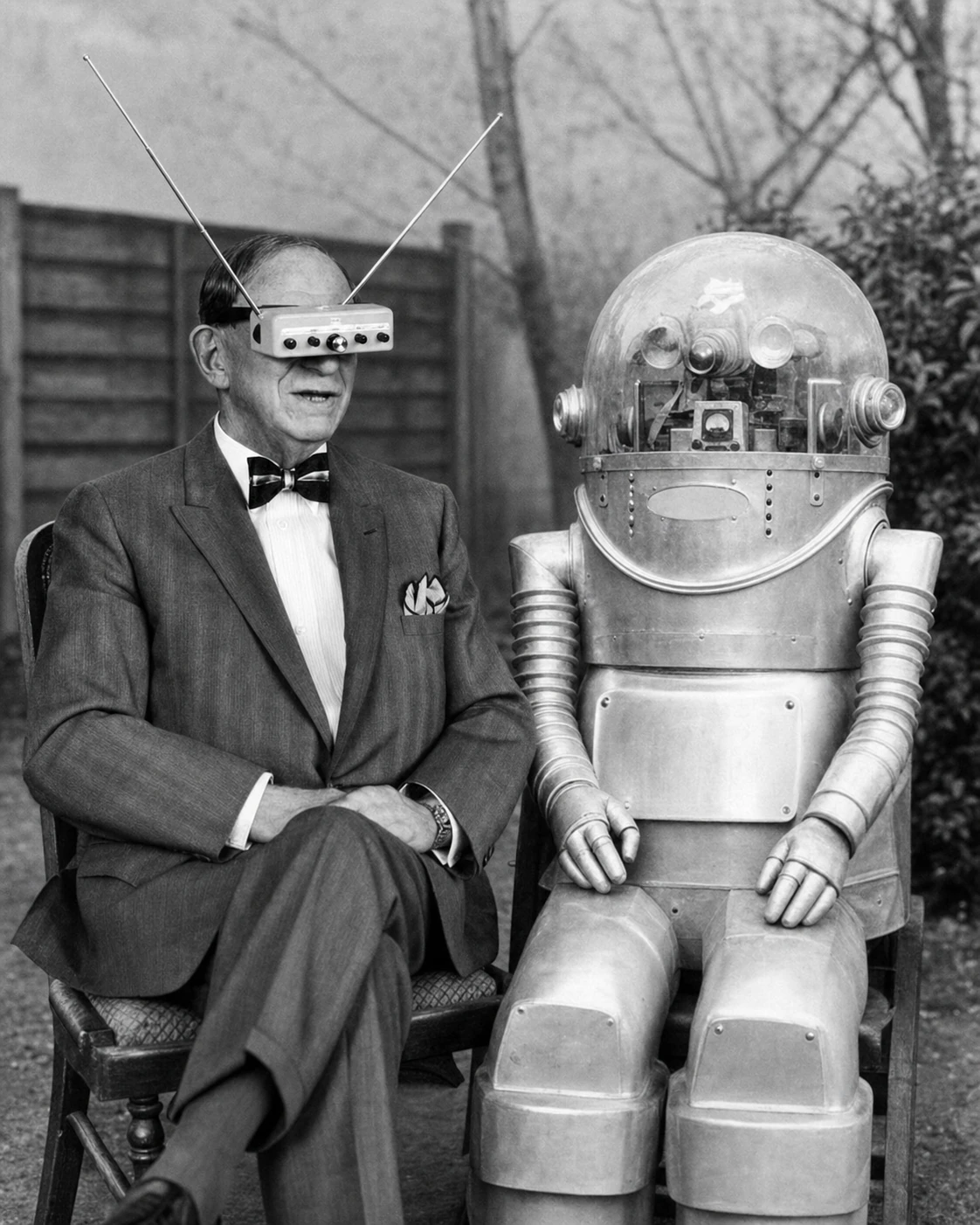 A vintage black-and-white photograph of a dapper gentleman wearing retro-futuristic electronic eyewear seated beside a humanoid robot in a garden.