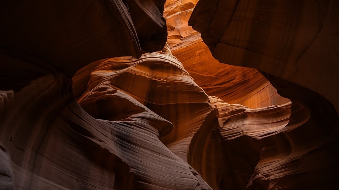 The image captures a stunning view inside a narrow slot canyon, likely Antelope Canyon. The walls are formed of swirling, undulating sandstone in shades of deep orange, reddish-brown, and lighter peach tones. The rock formations are smooth and organic, resembling flowing water or sculpted waves. The lighting is dramatic, with strong contrasts between the illuminated sections and the deep shadows, enhancing the texture and depth of the canyon walls. The composition is tight, focusing on the intricate patterns and shapes of the sandstone, creating a sense of enclosure and mystery. The overall impression is one of natural beauty, geological wonder, and the power of erosion over time.