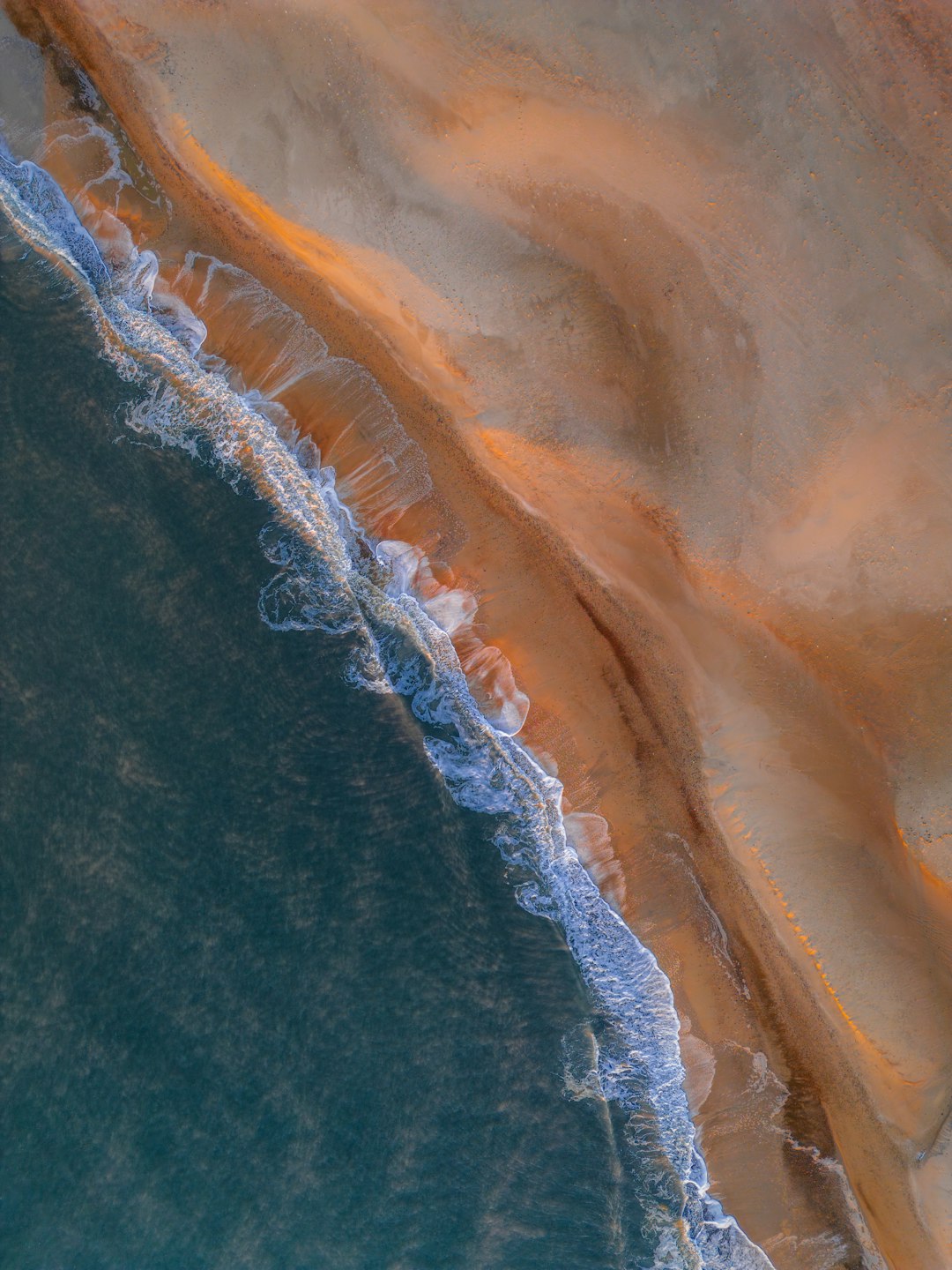 This is a high-angle aerial photograph of a beach and ocean. The image is dominated by the contrasting colors of the turquoise ocean and the golden-brown sand. The waves are visible as white foam breaking along the shoreline, creating a textured edge between the water and the sand. The sand appears to be a mix of light and darker shades, possibly indicating varying levels of moisture or different types of sediment. The composition is simple and focuses on the natural patterns and textures of the coastline. The overall impression is one of tranquility and natural beauty.