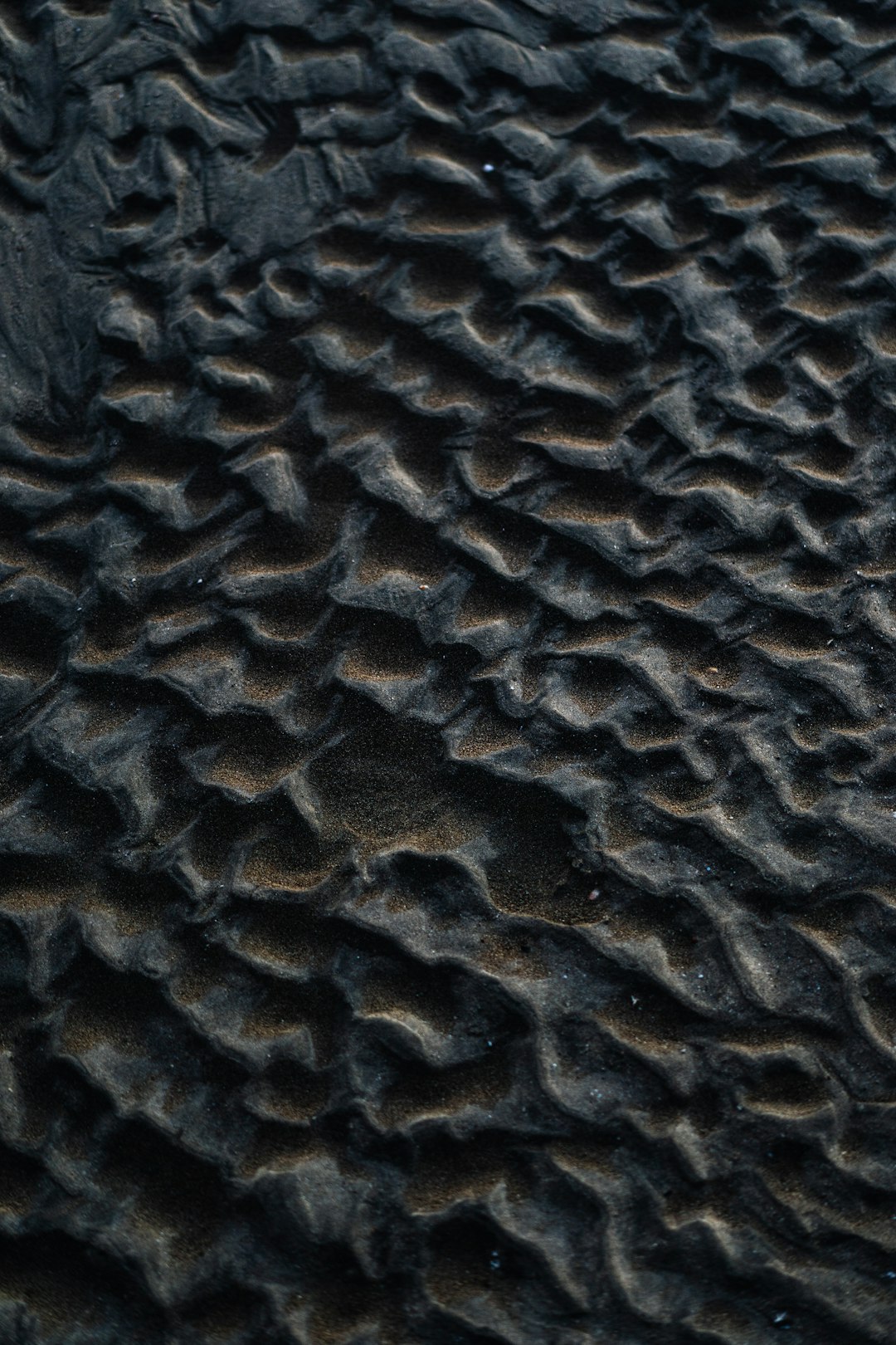 The image is a close-up, top-down view of dark, rippled sand. The sand forms a textured pattern of small, wave-like ridges and valleys, creating a sense of depth and movement. The color palette is dominated by dark tones, with shades of gray and black, and hints of golden or tan where the ridges catch the light. The overall effect is abstract and organic, resembling a miniature landscape or a topographic map. The lighting is soft and diffused, emphasizing the texture of the sand and creating subtle shadows.