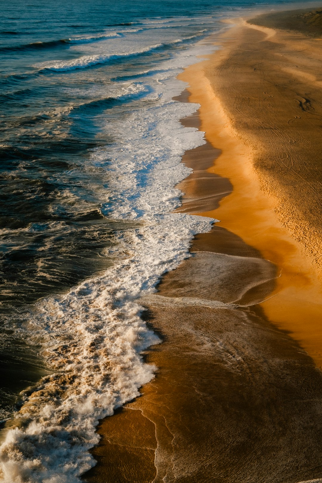 This is a high-angle, aerial view of a beach and the ocean. The image is vertically oriented, emphasizing the long stretch of coastline. 

The left side of the image is dominated by the ocean, with waves rolling towards the shore. The water is a deep blue, with white foam and crests marking the breaking waves. The waves are creating a textured pattern as they approach the beach.

The right side of the image shows the sandy beach. The sand is a warm, golden-brown color, and it's marked with intricate patterns created by the receding waves and the flow of water. These patterns resemble small rivers or channels winding through the sand.

The contrast between the dark blue ocean and the golden sand is striking. The image captures the dynamic interplay between the water and the land, creating a sense of movement and tranquility. The lighting appears to be soft and diffused, enhancing the natural colors and textures of the scene.