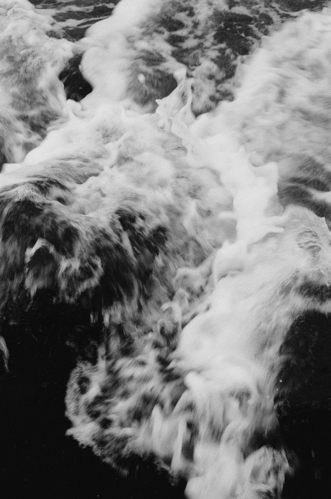 This is a dramatic black and white photograph of turbulent water. The image is filled with swirling, frothy white foam against a dark, almost black background. The water appears to be rushing or cascading, creating a sense of motion and energy. The foam is dense and textured, with a chaotic pattern of peaks and valleys. The contrast between the white foam and the dark water is very strong, emphasizing the dynamic nature of the scene. The overall impression is one of power, movement, and a slightly ominous beauty.