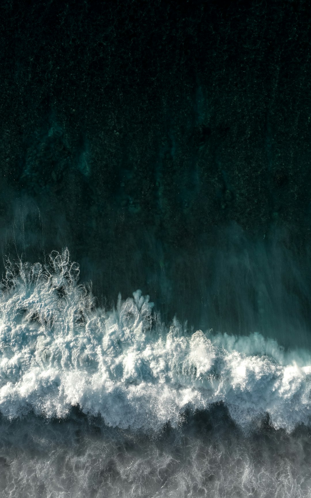 The image depicts a turbulent ocean scene, likely a wave breaking or a strong current. The dominant color is a deep, dark teal, representing the water. The lower portion of the image is filled with white, frothy foam and churning water, indicating the crest of a wave or the impact of water against a surface. The foam appears almost like wisps of smoke, creating a dynamic and chaotic texture. Above the foam, the water transitions into a darker, smoother teal, suggesting the deeper parts of the ocean. The overall impression is one of power, movement, and the raw energy of the sea. The image has a slightly abstract quality due to the way the foam is rendered, resembling a painterly effect.