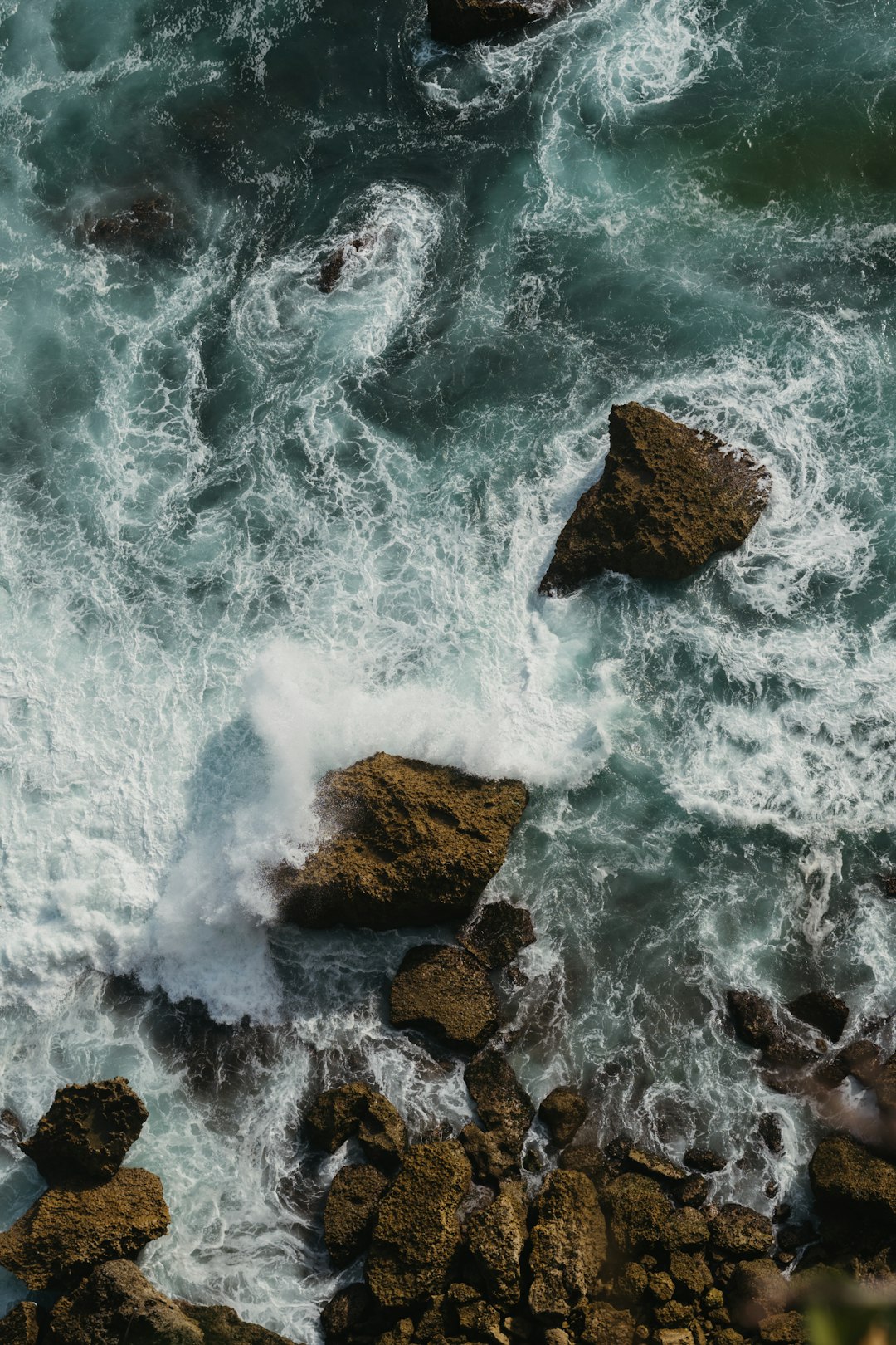 This is a high-angle, aerial view of a rocky coastline with crashing waves. The water is a deep teal color, swirling with white foam and turbulence as it collides with the dark, jagged rocks. The rocks are various sizes and shapes, some partially submerged, and others standing tall against the force of the waves. The waves are dynamic, creating whitecaps and splashes as they break around the rocks. The overall impression is one of raw natural power and the constant interplay between land and sea.