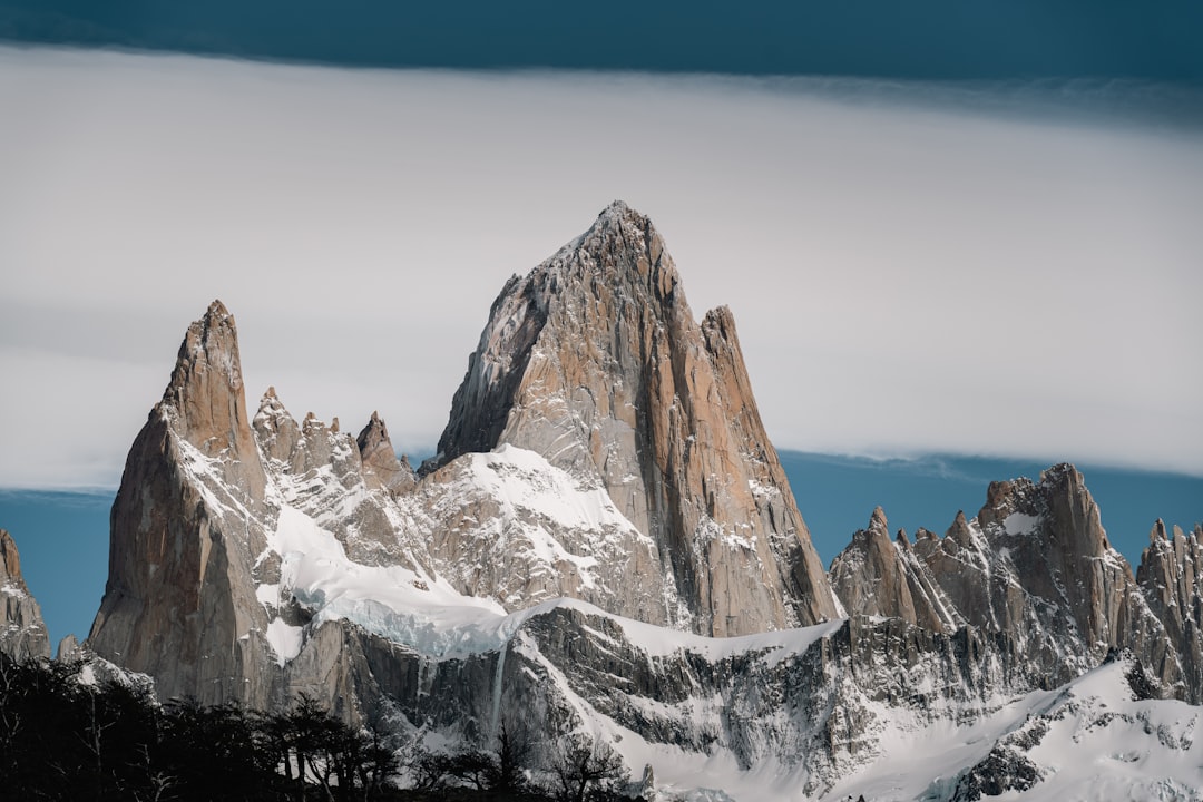 The image showcases a dramatic mountain range, dominated by a towering, jagged peak covered in snow and ice. The mountain is a mix of exposed rock and snow-covered areas, with steep cliffs and ridges. The peak is the focal point, rising sharply from the surrounding landscape. The lower slopes of the mountain are covered in snow, with patches of exposed rock. A few dark, silhouetted trees are visible at the base of the mountain. The sky is a muted blue-grey, with a layer of clouds partially obscuring the peak. The overall impression is one of rugged beauty and remote wilderness.