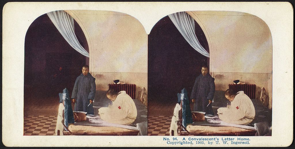 Stereoscopic photograph of a man writing a letter while a woman in a red cross uniform kneels beside him.