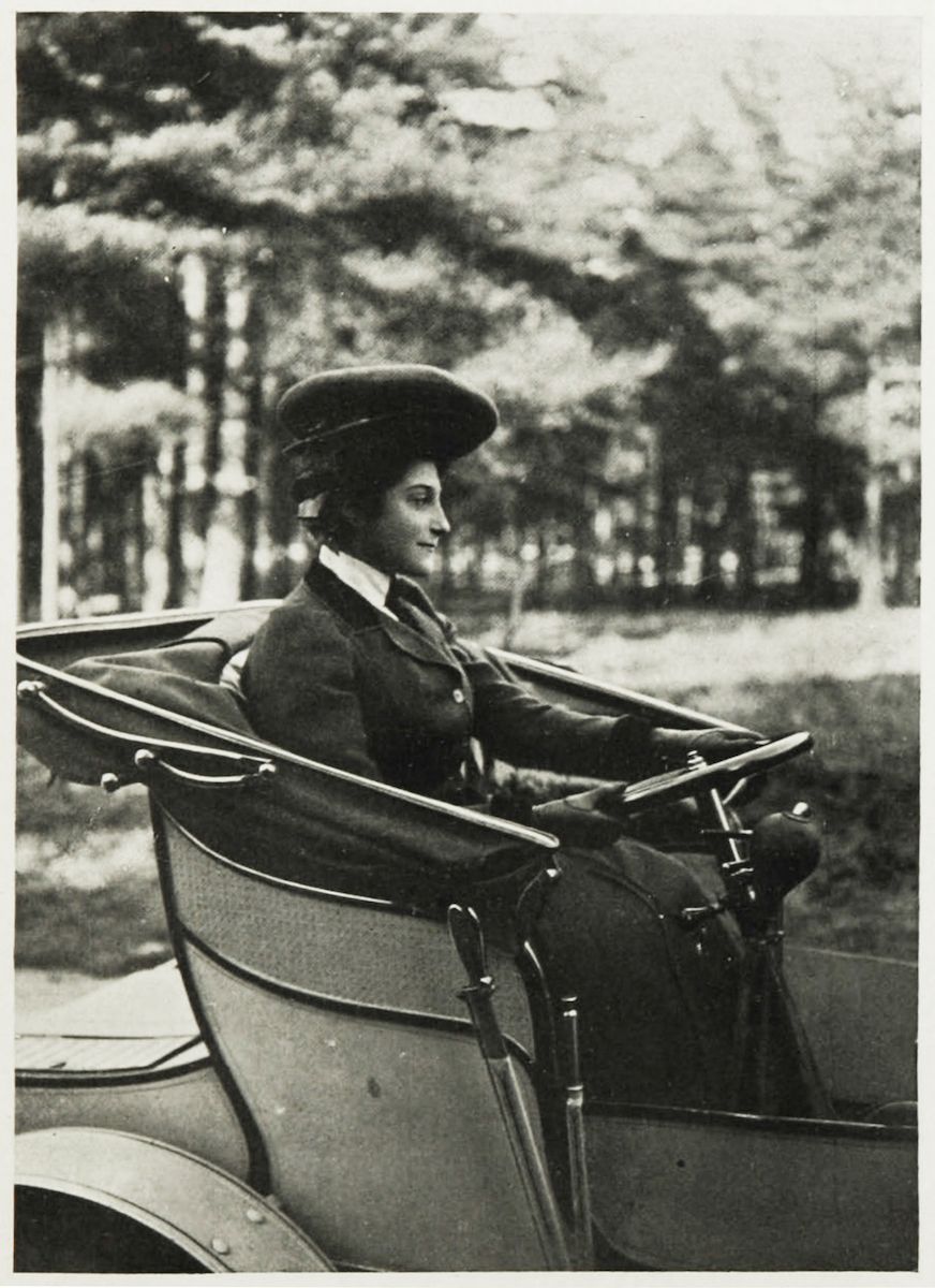 A vintage black and white photograph of a woman driving an early automobile.