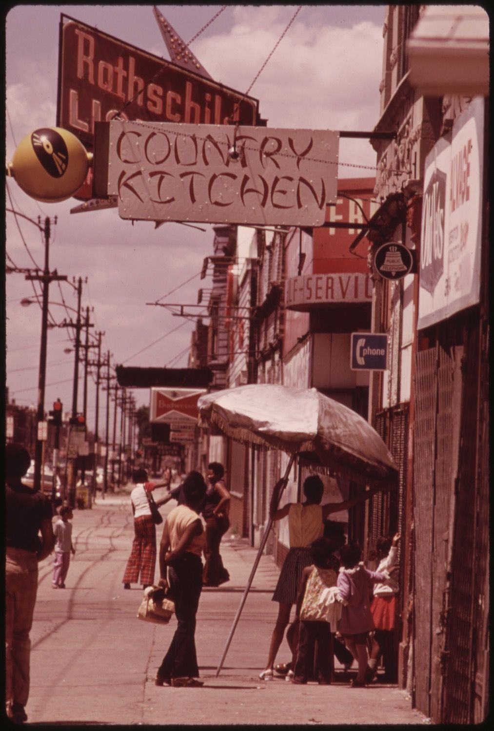 A vintage street scene with a sign for Rothschild's Country Kitchen, people walking on the sidewalk, and a makeshift canopy.