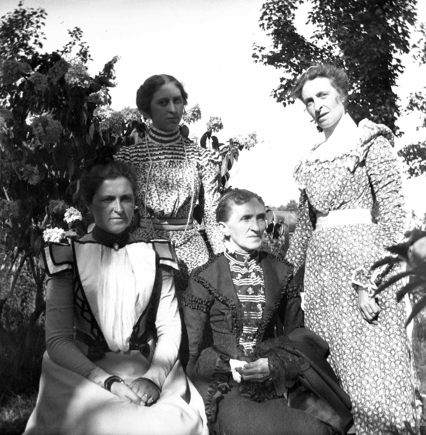 A black and white photograph of five women in long dresses, likely taken outdoors in a garden or natural setting.