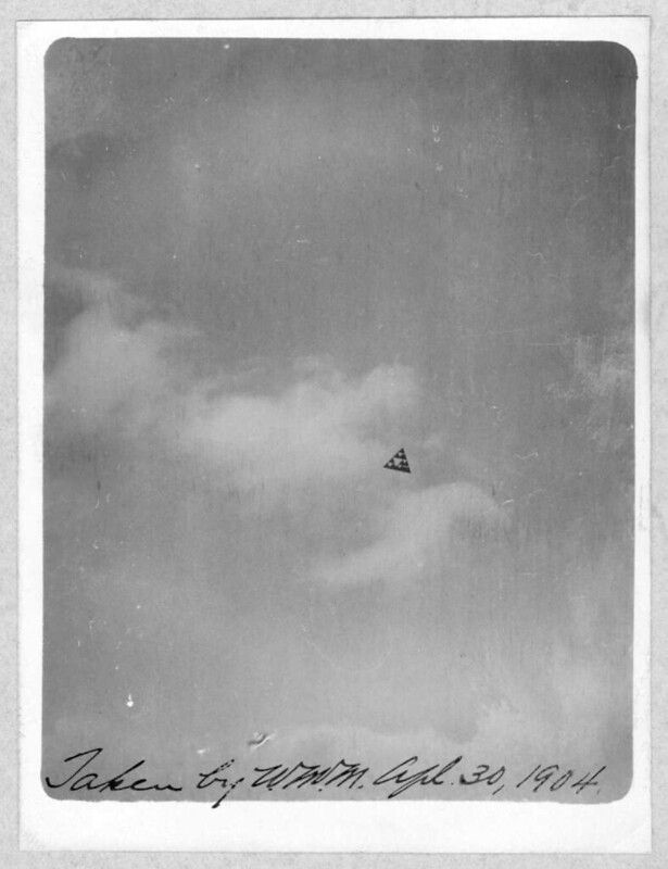 A black and white photograph of an early biplane in flight against a hazy sky, with handwritten text at the bottom reading 'Taken by W.M. July 30, 1904.'