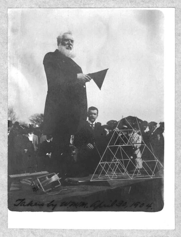 A vintage black and white photograph from 1904 showing a tall man with a long white beard standing next to a large, geometric structure resembling a pyramid made of metal wires. A crowd of people are gathered around, observing the scene.