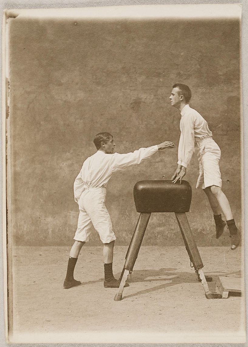 Two young boys in vintage athletic wear performing a gymnastic exercise over a wooden horse.