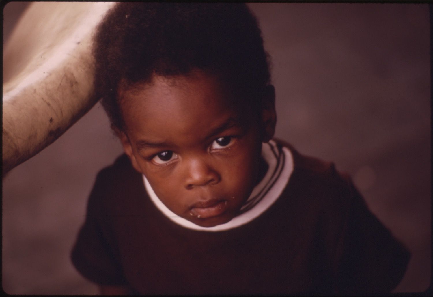 Close-up portrait of a young Black child with a serious expression, wearing a dark sweater with a white collar.