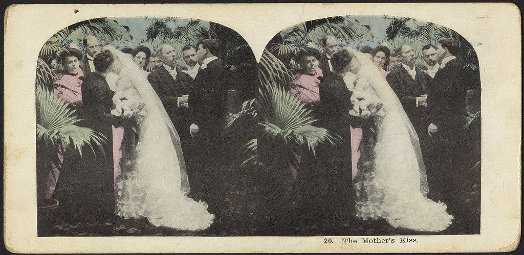 Stereoscopic card titled "The Mother's Kiss" depicting a woman in a wedding dress being kissed by a man, surrounded by a group of people in a lush outdoor setting.
