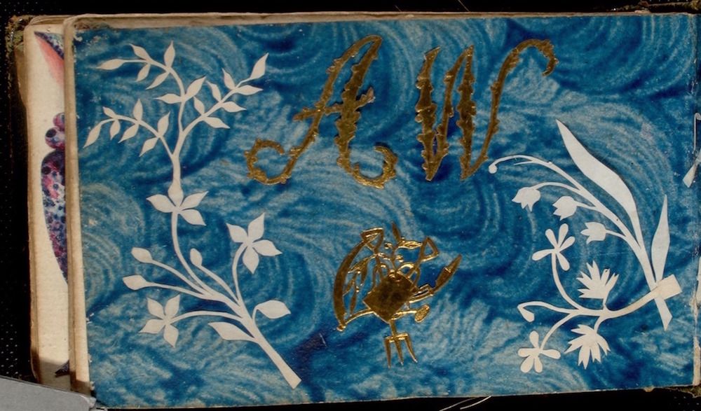 Close-up of a vintage book cover with a blue marbled background, white floral branches, gold lettering 'A.M.C.', and a gold crab.