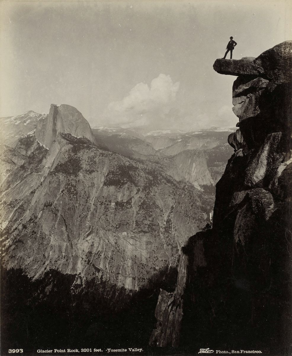 A vintage black and white photograph of Yosemite Valley, showing a person standing on Glacier Point Rock overlooking the vast landscape of granite cliffs and forests. Half Dome is visible in the distance.