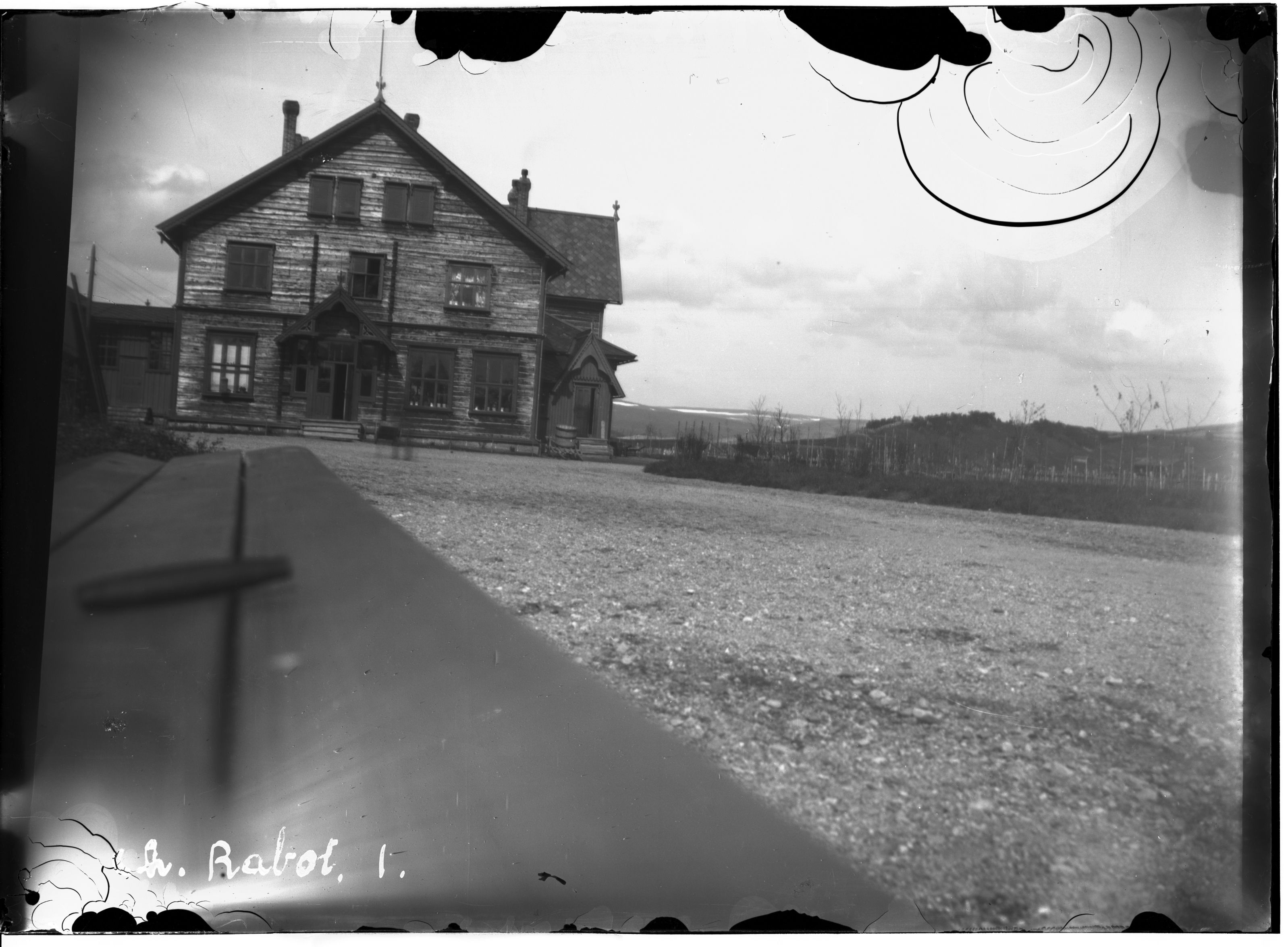 A black and white photograph of a large, two-story house with a steeply pitched roof and a porch. The house is surrounded by a yard with a fence, and there are trees in the background. The sky is overcast, and the ground is covered in gravel.