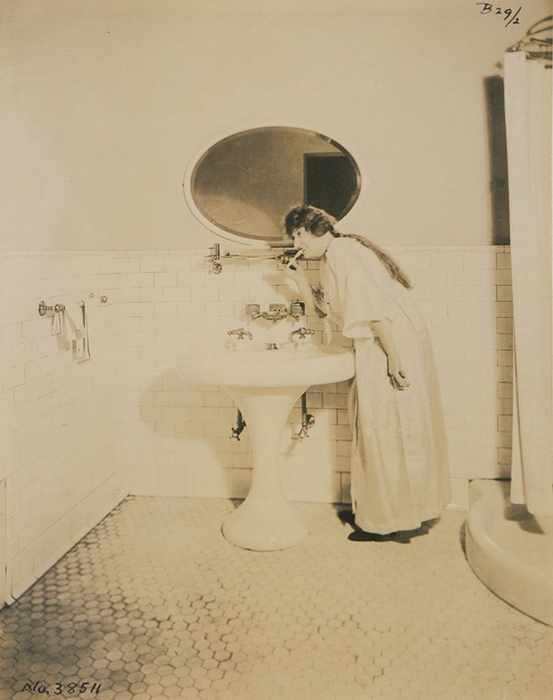 Vintage black and white photograph of a woman washing her hands in a tiled bathroom.