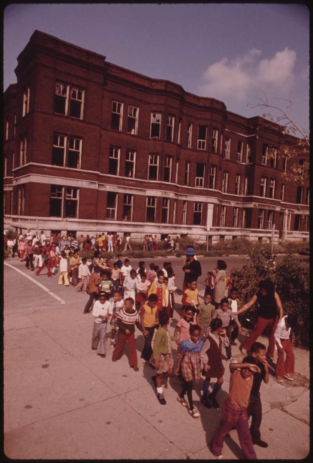 A large group of children and adults walking towards the camera in front of a three-story red brick building.