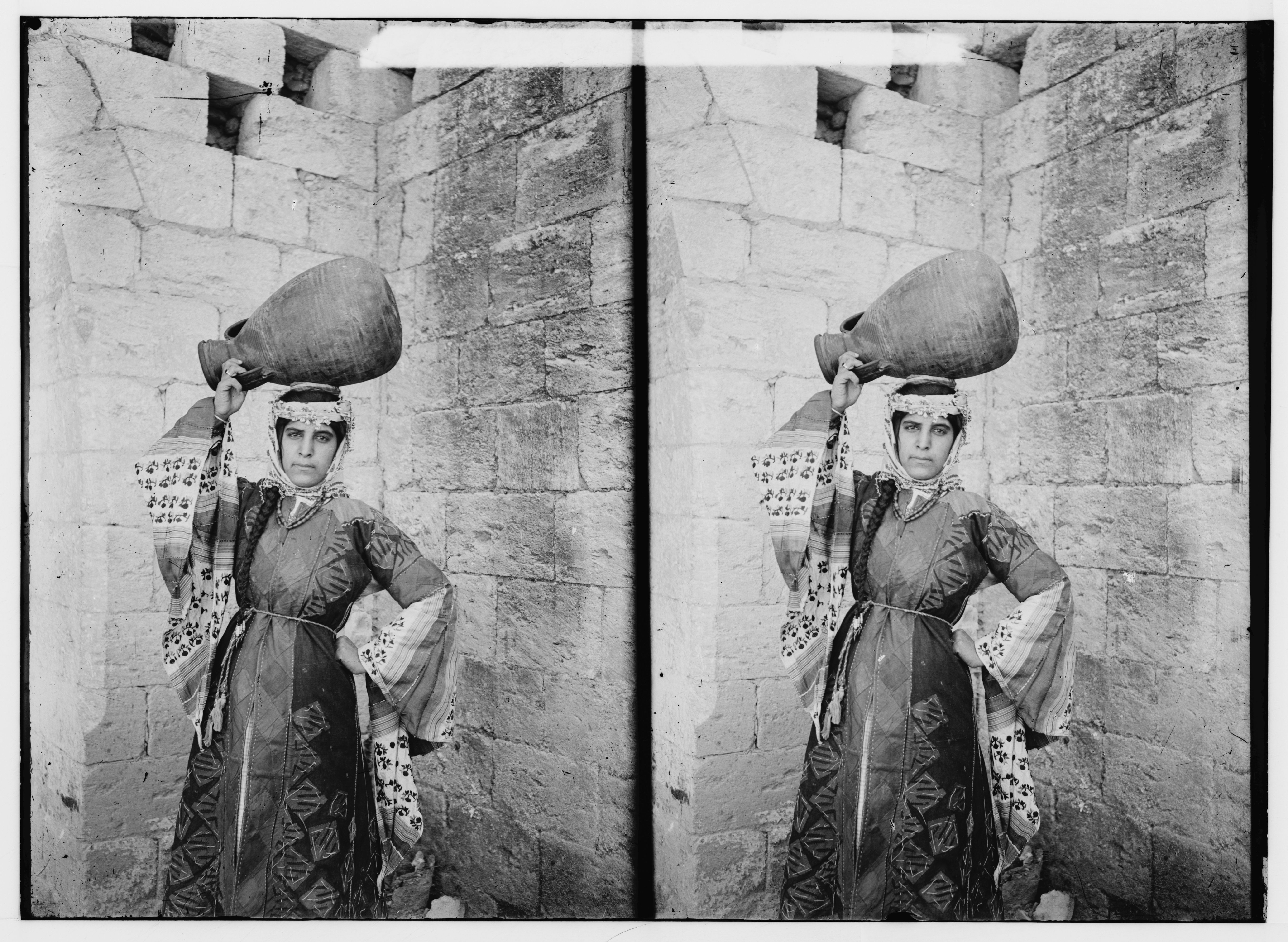 Stereoscopic photograph of a woman balancing a jar on her head against a stone wall.